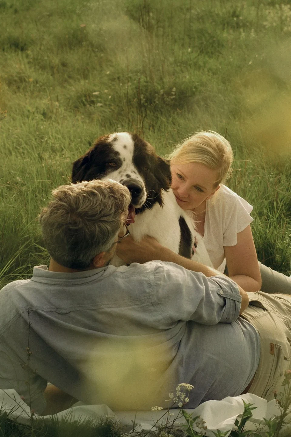 Couple sitting in a meadow with the woman leaning against their dog
